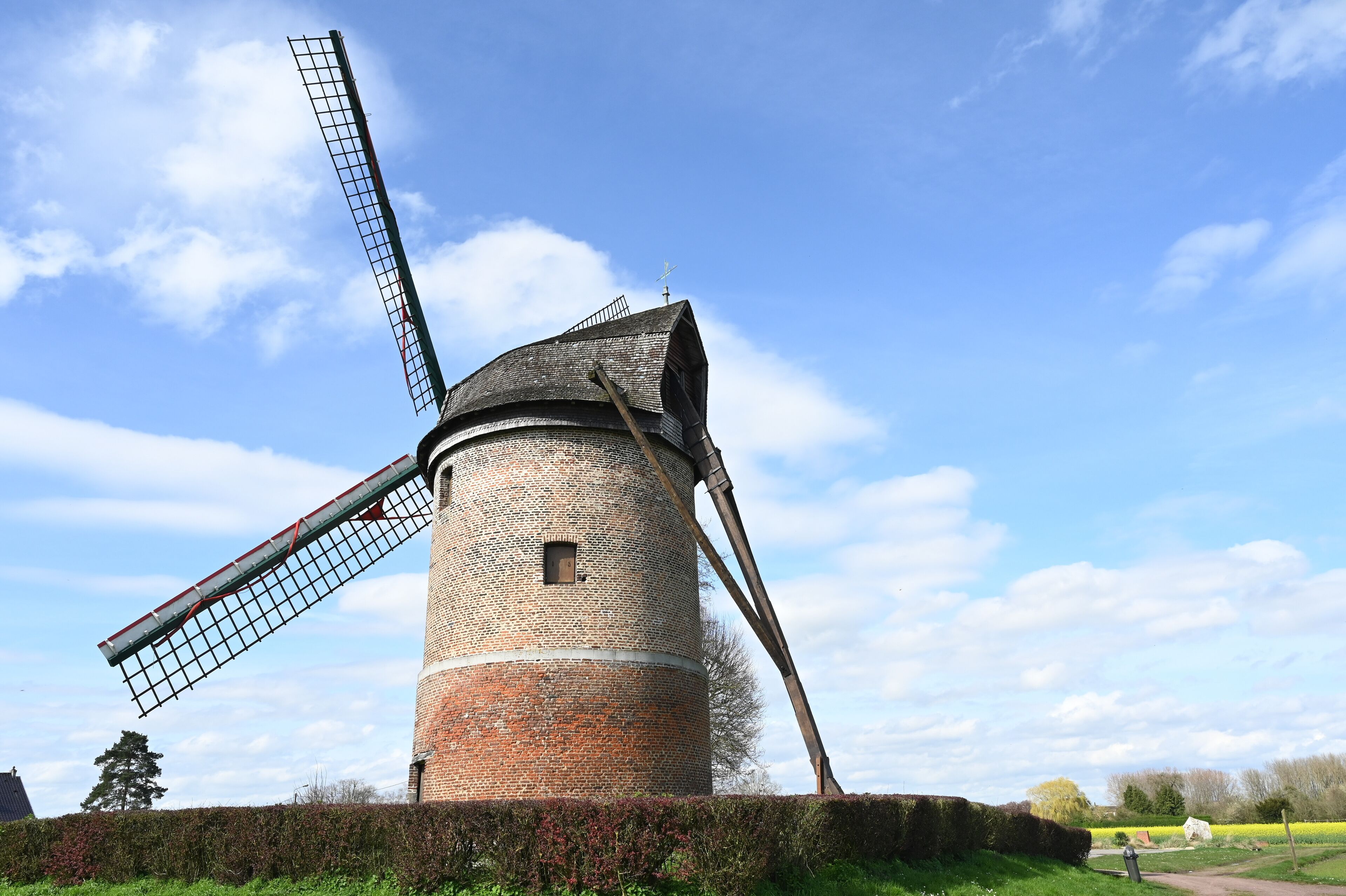 A traditional windmill with a large sail and a brick base, surrounded by greenery and a clear sky.