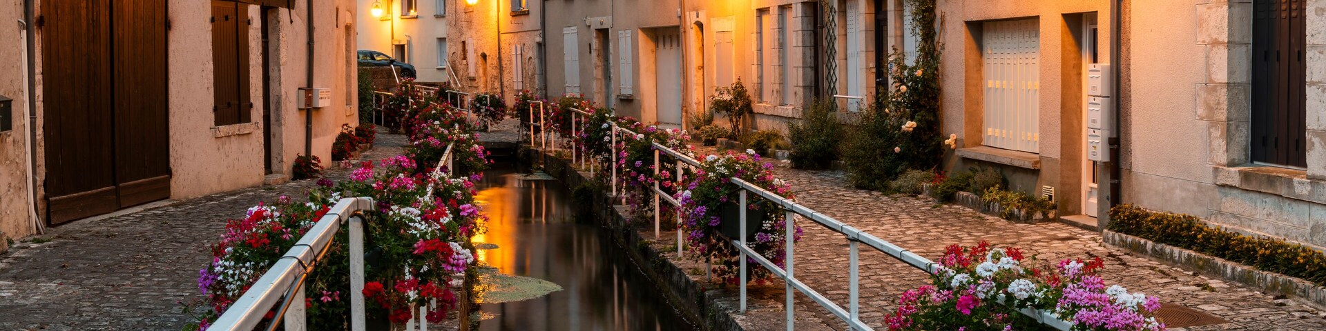 Um canal estreito ladeado por edifícios históricos, com grades adornadas por flores e iluminação acolhedora.