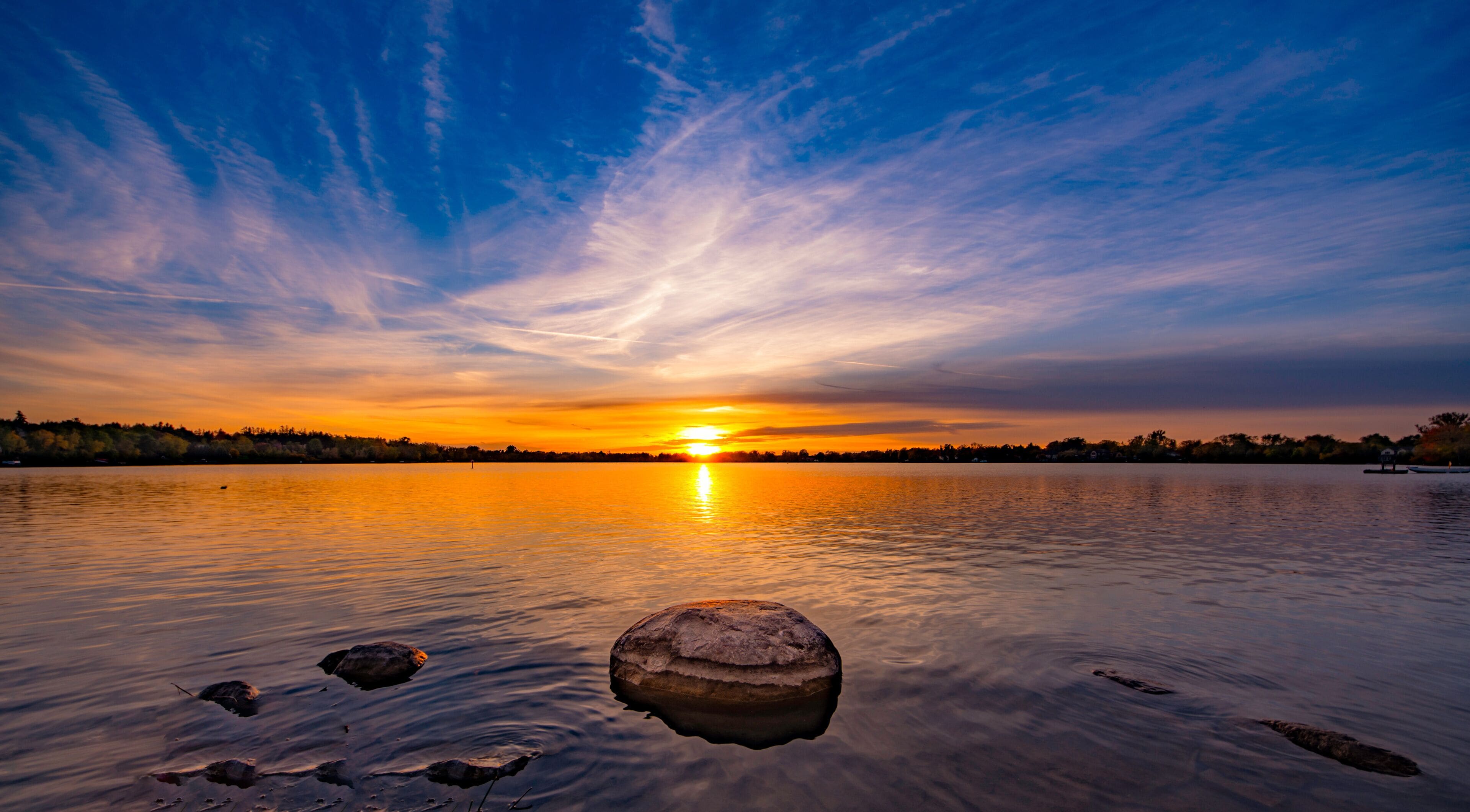 A sunset over a calm lake with a rock in the foreground and a boat on the right.
