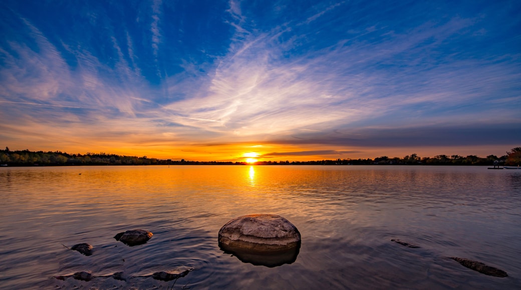 A sunset over a calm lake with a rock in the foreground and a boat on the right.
