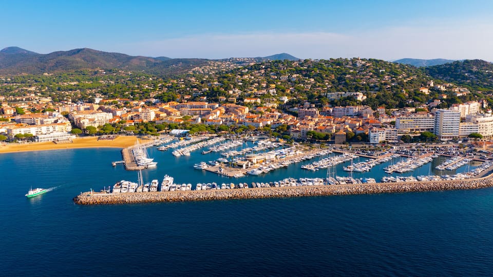 A coastal town with a marina, sandy beach, and hilly background.