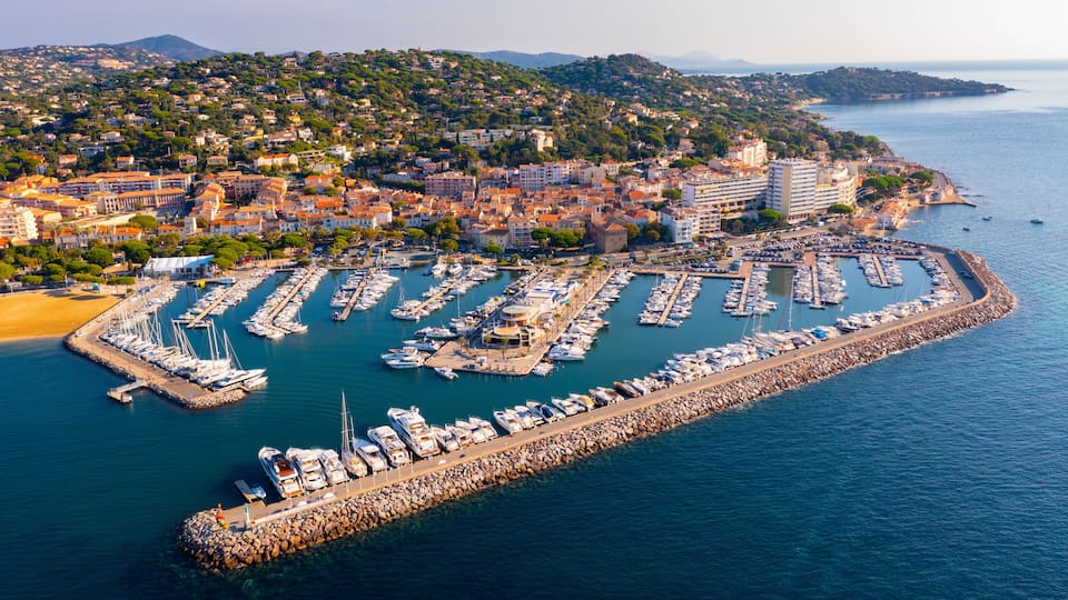 A coastal marina with numerous boats docked, a rocky breakwater, and a hillside town in the background.