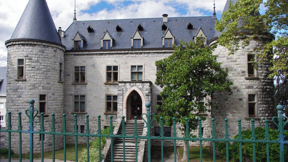 A historic stone castle with a conical tower, arched windows, and a green metal fence.