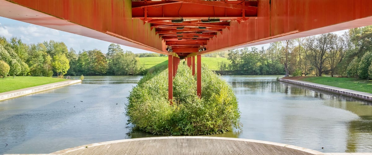 A wooden deck under a red metal structure over a calm river with greenery and trees in the background.