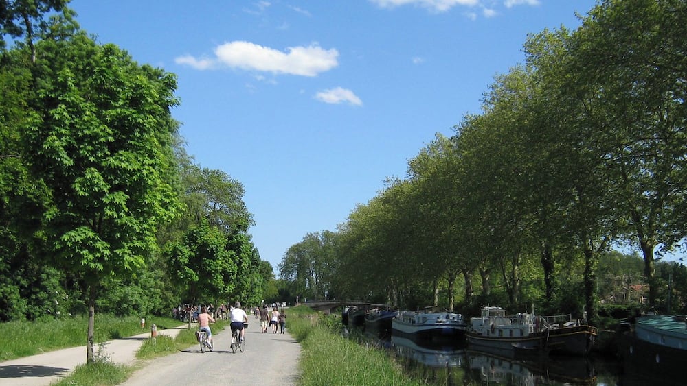 Un canal bordé de bateaux, fréquenté par des cyclistes et entouré d’une végétation luxuriante.
