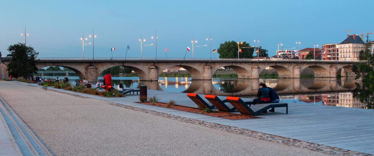 A stone bridge over a calm river, benches along the riverbank, and flags on the bridge.