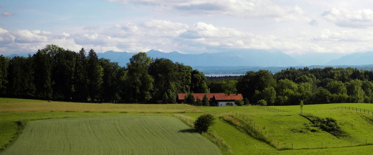 A rural landscape with green fields, a red-roofed house, and a forested area with mountains in the background.
