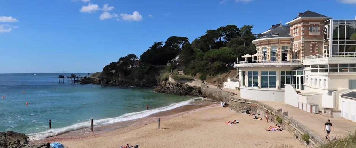 A coastal beach with clear skies, a rocky outcrop, and a large building with a balcony overlooking the sea.