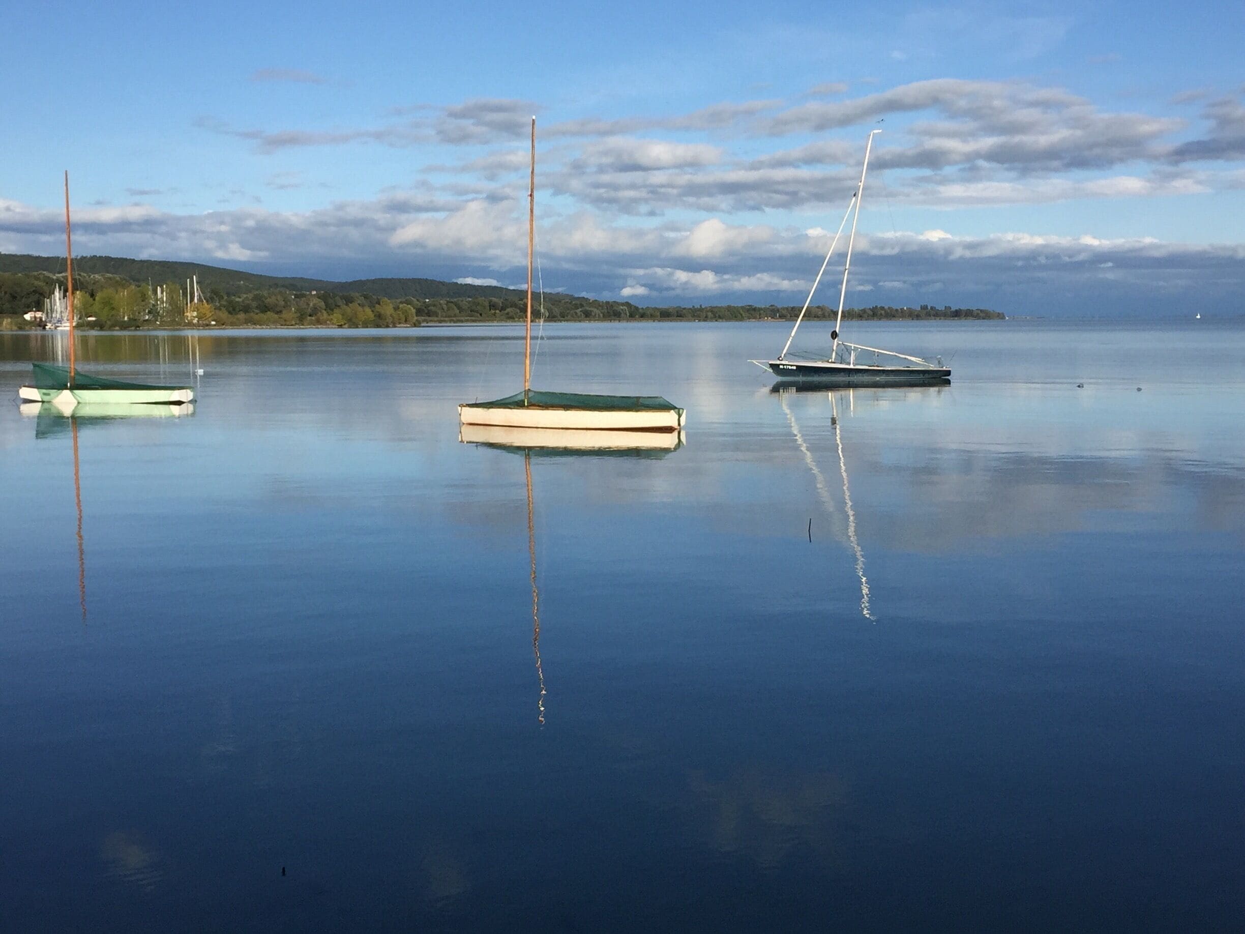 Calm water with three sailboats anchored near a shoreline with trees and hills in the background.
