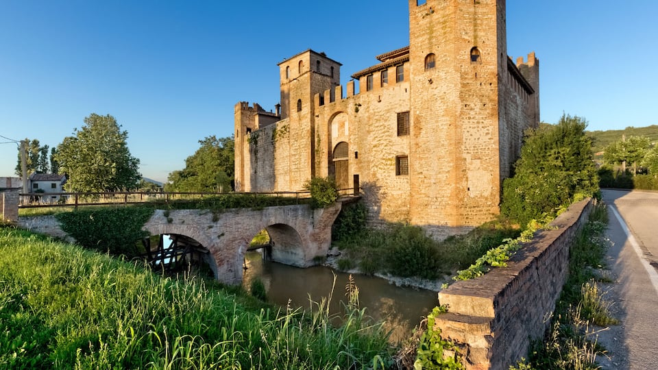 A historic stone castle with a bridge over a river, surrounded by greenery and a clear sky.