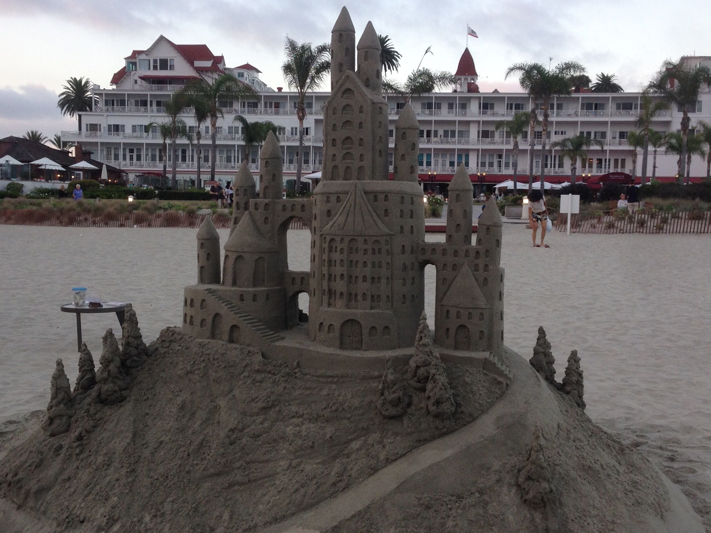 A sandcastle with towers and a bridge, surrounded by palm trees and a beach.