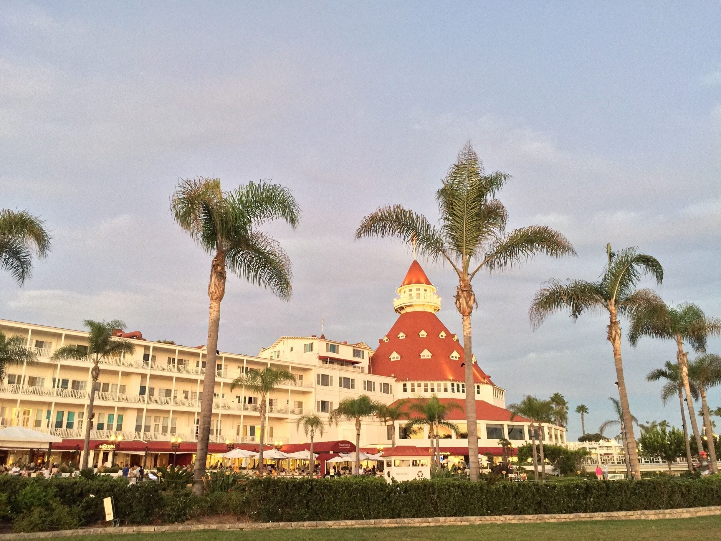 A hotel with a distinctive red roof and a white building with balconies. Palm trees line the front of the hotel.