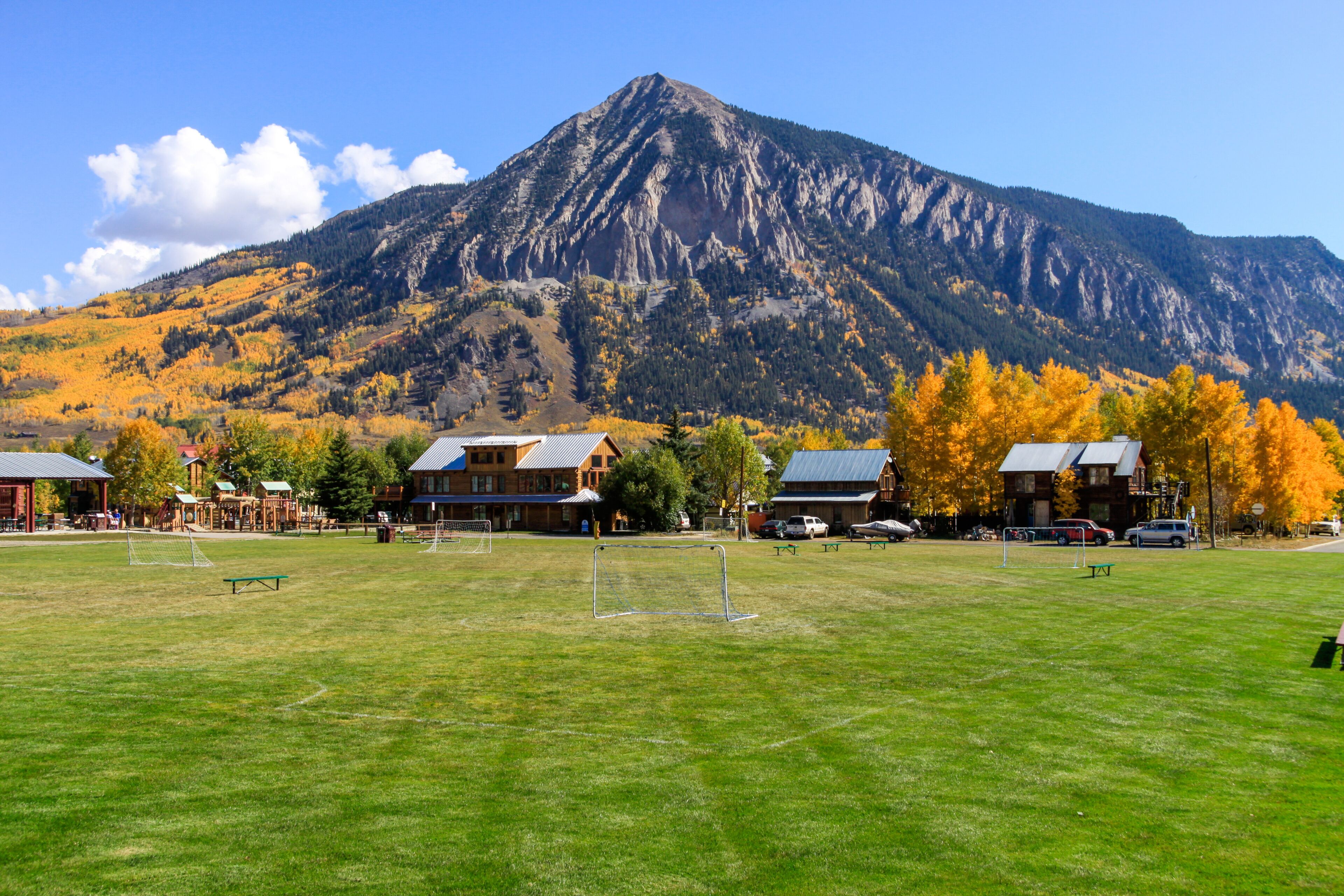 A grassy field with a soccer goal, a wooden cabin, and a mountain in the background.