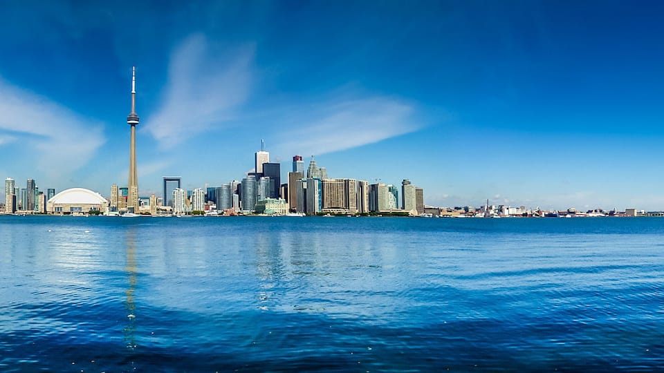 A city skyline with a prominent tower, a body of water in the foreground, and a clear blue sky.