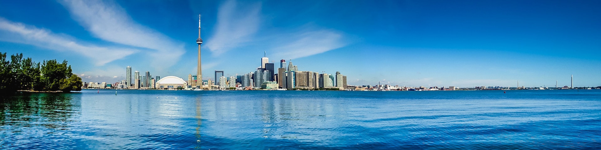 Eine Stadtlandschaft mit einem markanten Turm, einem Wasserlauf im Vordergrund und blauem, wolkenlosem Himmel.