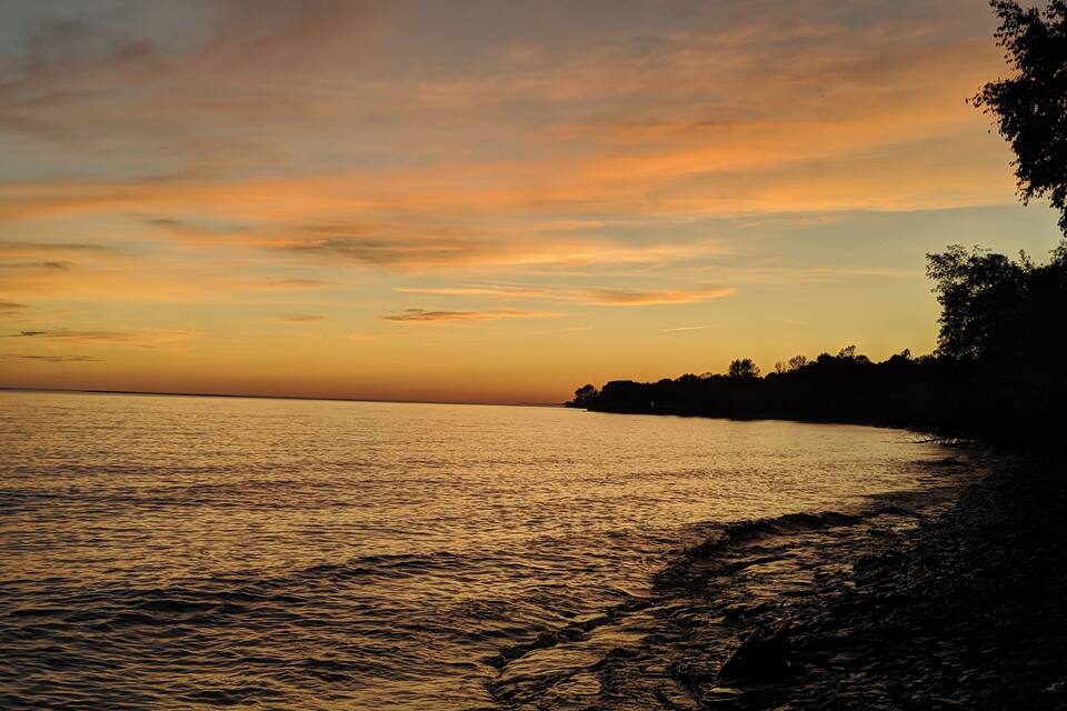 A sunset over a calm sea with a rocky shoreline and silhouetted trees.