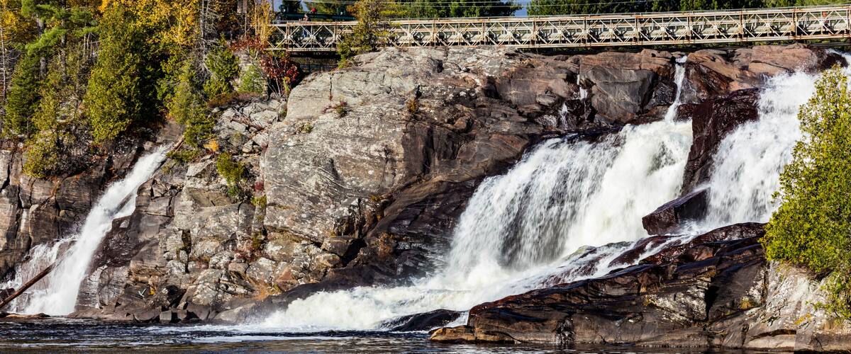 Una cascada en varios niveles que se precipita sobre terreno rocoso, con un puente al fondo.