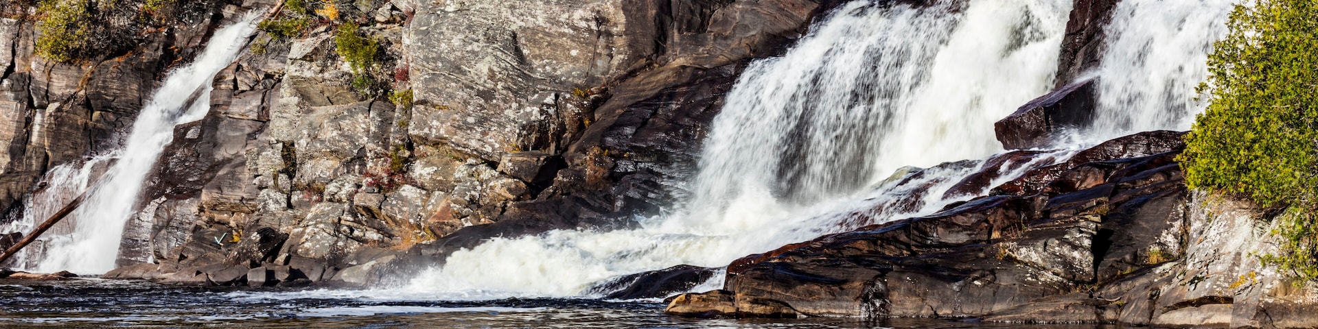 Uma cascata em vários níveis deságua sobre terreno rochoso, com uma ponte ao fundo.
