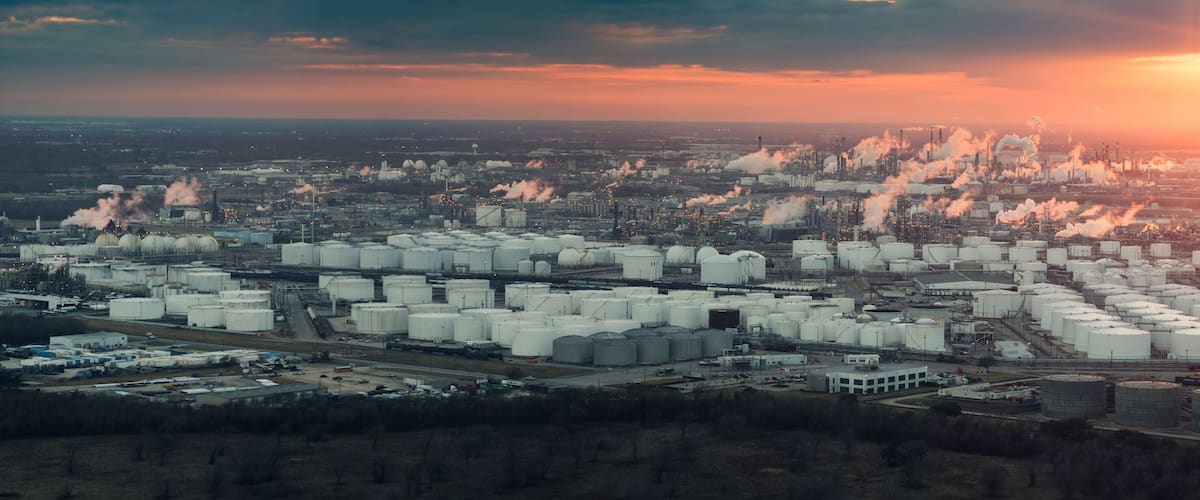 An industrial complex with multiple storage tanks and smokestacks emitting smoke, set against a sunset sky.