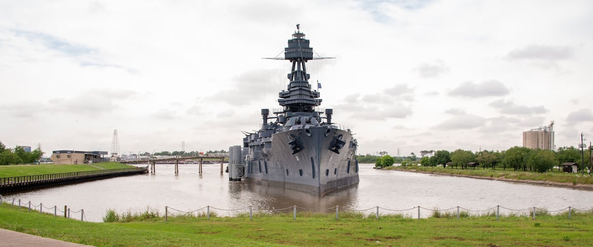 Een groot marineschip afgemeerd in een rivier, omringd door begroeide oevers en een brug op de achtergrond.