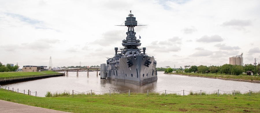 A large naval ship docked in a river, surrounded by grassy banks and a bridge in the background.