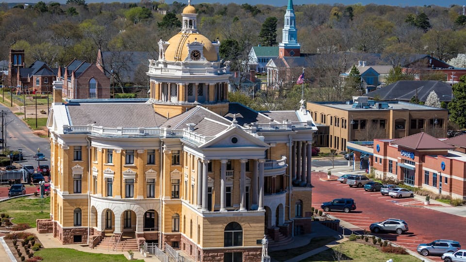 A historic building with a yellow dome and columns, surrounded by a parking lot and other buildings.