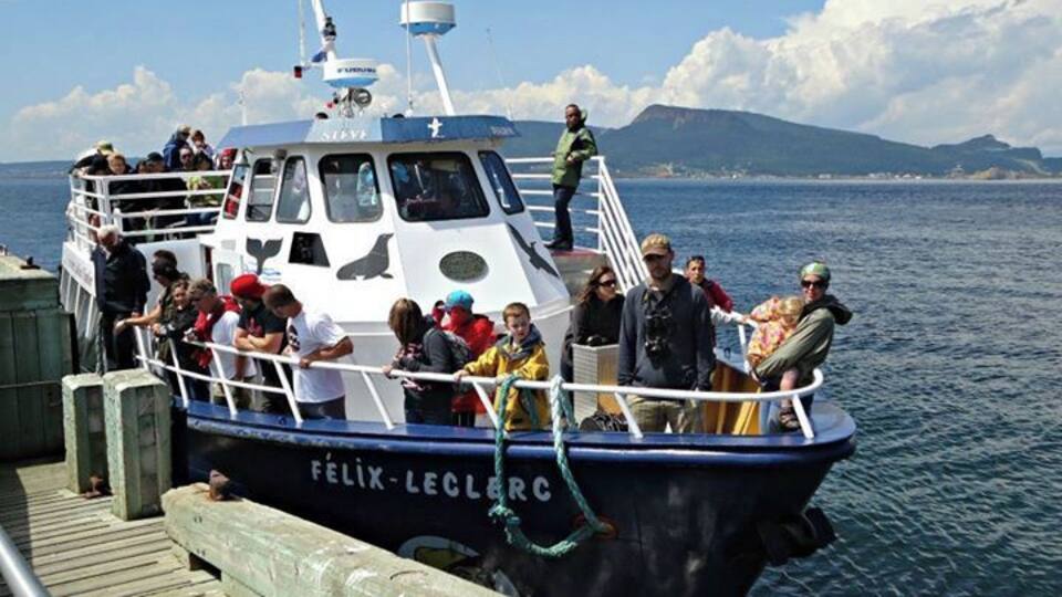 A boat named 'Félix-Leclercq' docked at a pier with people on board and a mountainous landscape in the background.