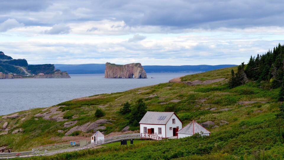 A coastal landscape with a white house, a wooden bridge, and a large rock formation in the distance.