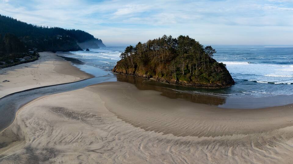 A coastal landscape with a sandy beach, a rocky outcrop, and a forested hillside.