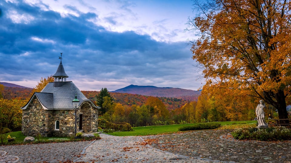A stone church with a pointed steeple, surrounded by autumn foliage and mountains.