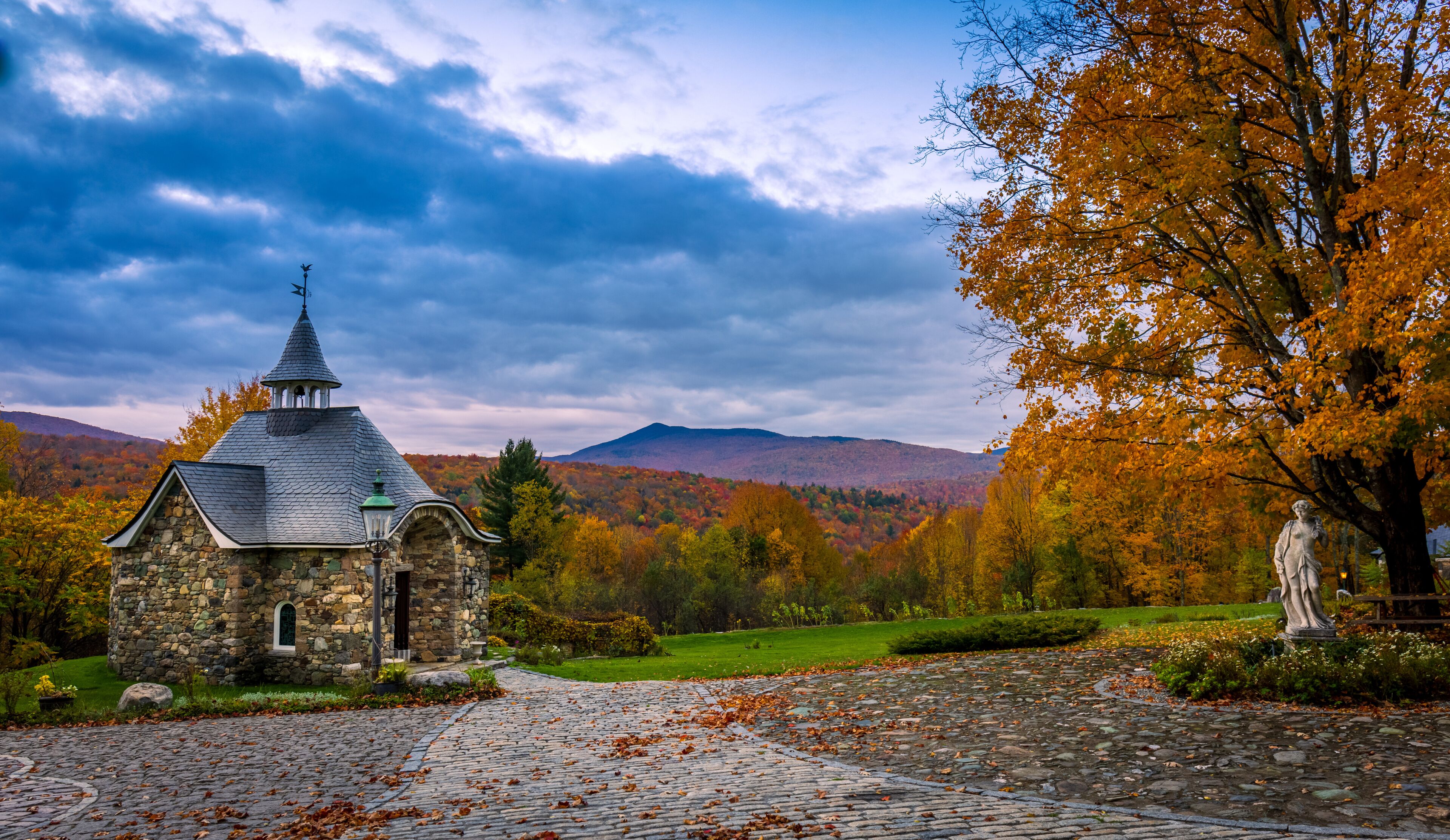 A stone church with a pointed steeple, surrounded by autumn foliage and mountains.