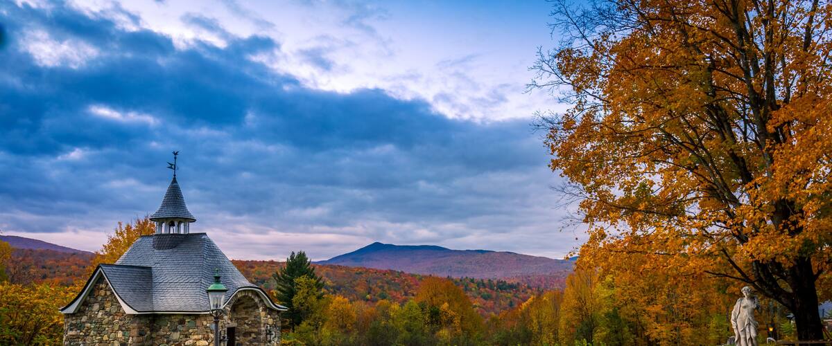 A stone church with a pointed steeple, surrounded by autumn foliage and mountains.