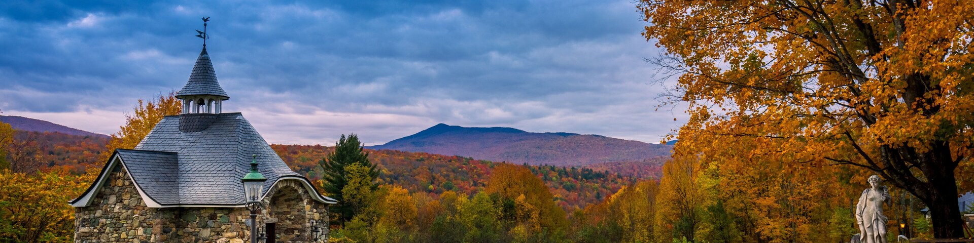 A stone church with a pointed steeple, surrounded by autumn foliage and mountains.