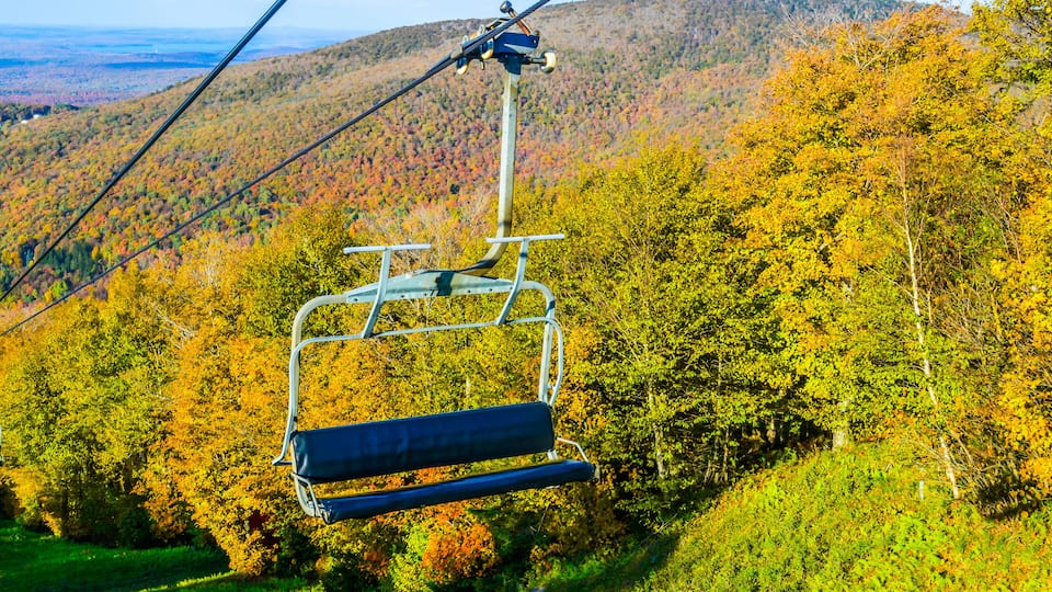 A cable car chair suspended over a forested mountain with autumn foliage.