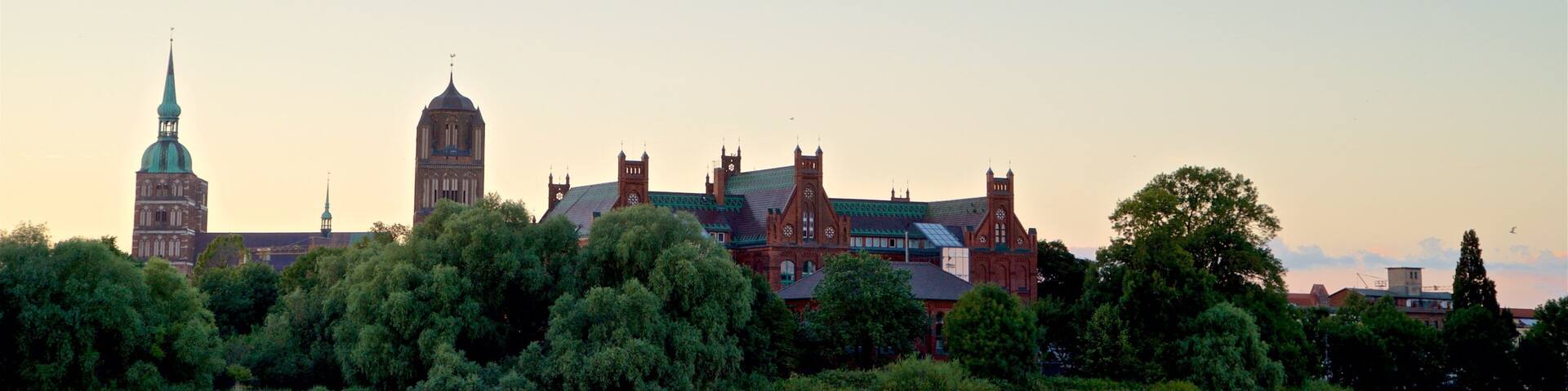 A historic building complex with a prominent clock tower and a green-domed church by a river.