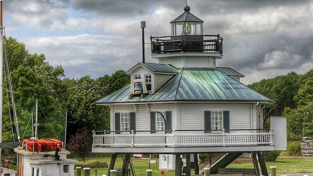 A lighthouse on stilts with a green roof, surrounded by water and greenery.