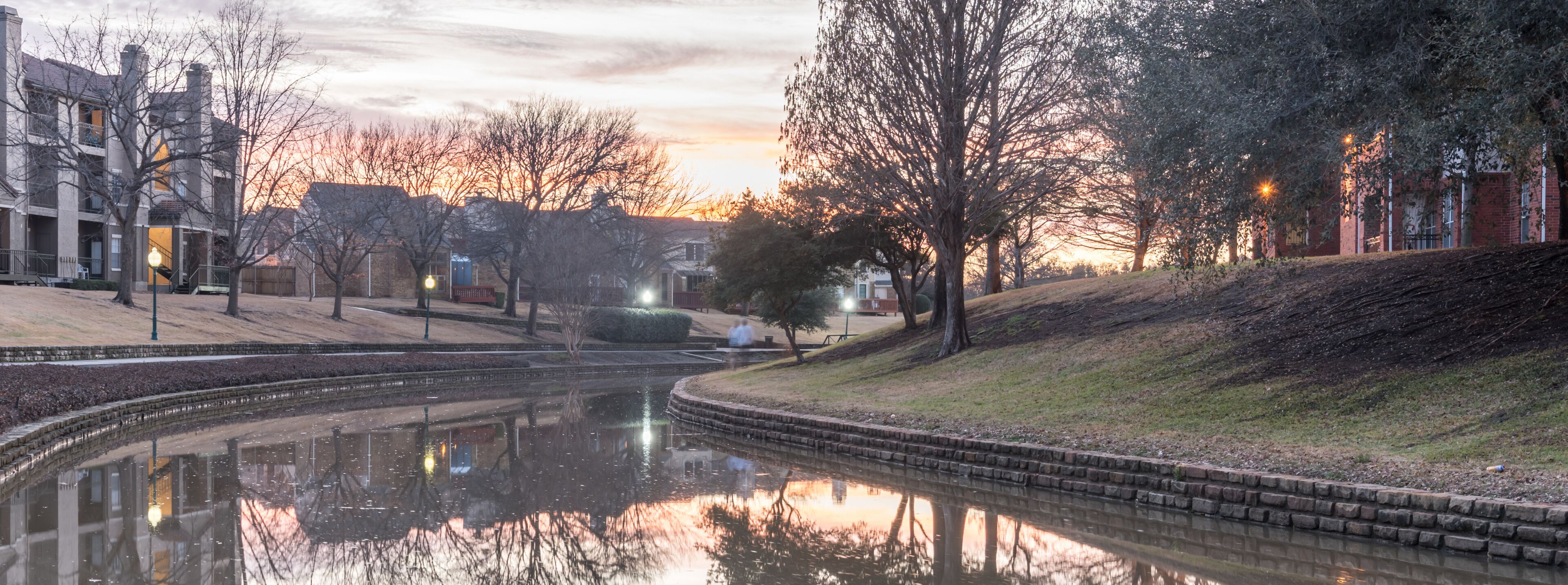A calm river reflecting buildings and trees, surrounded by a park with benches and a brick building.