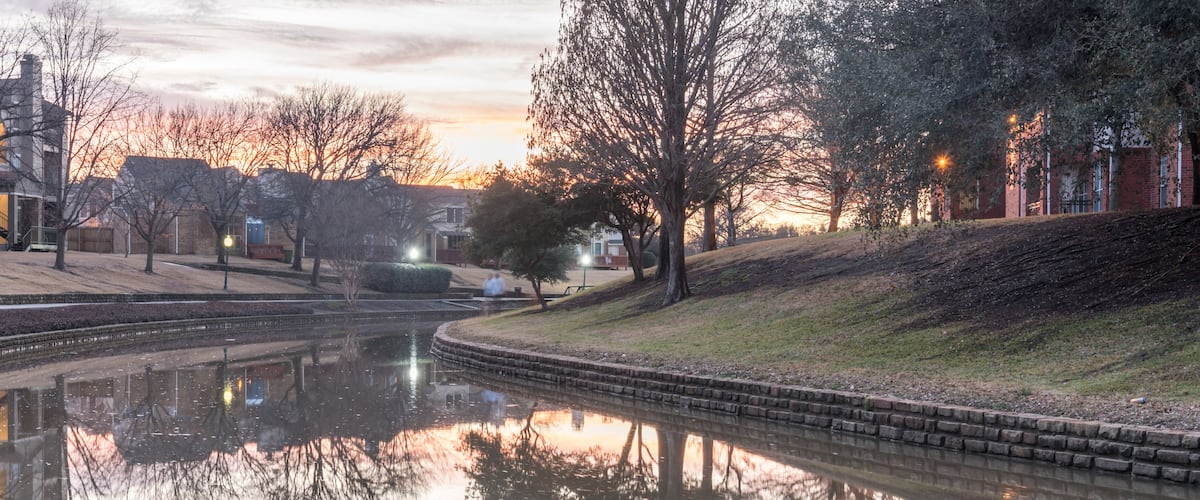 A calm river reflecting buildings and trees, surrounded by a park with benches and a brick building.