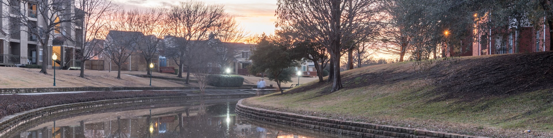 A calm river reflecting buildings and trees, surrounded by a park with benches and a brick building.