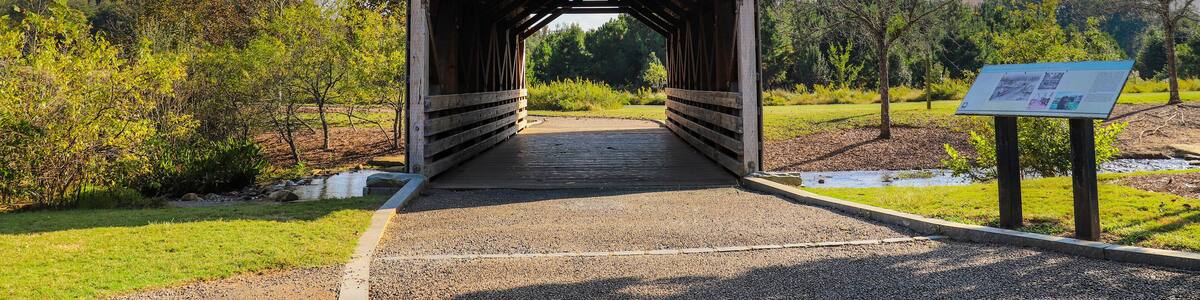 a shot of a wooden covered bridge over a river surrounded by lush green and autumn colored trees with lush green grass, blue sky and clouds at sunset at Garrard Landing Park in Alpharetta Georgia USA