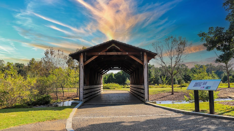 A covered bridge with a gravel path leading to it, surrounded by trees and a grassy area.