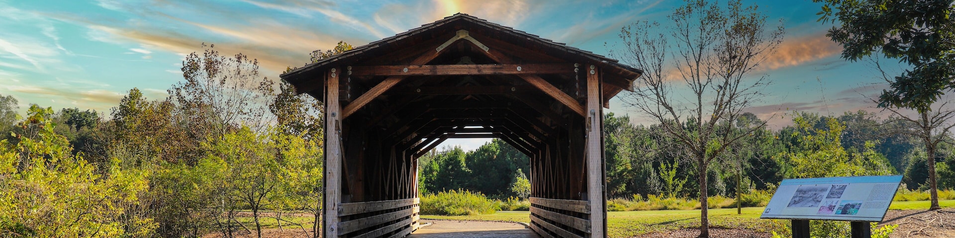 a shot of a wooden covered bridge over a river surrounded by lush green and autumn colored trees with lush green grass, blue sky and clouds at sunset at Garrard Landing Park in Alpharetta Georgia USA