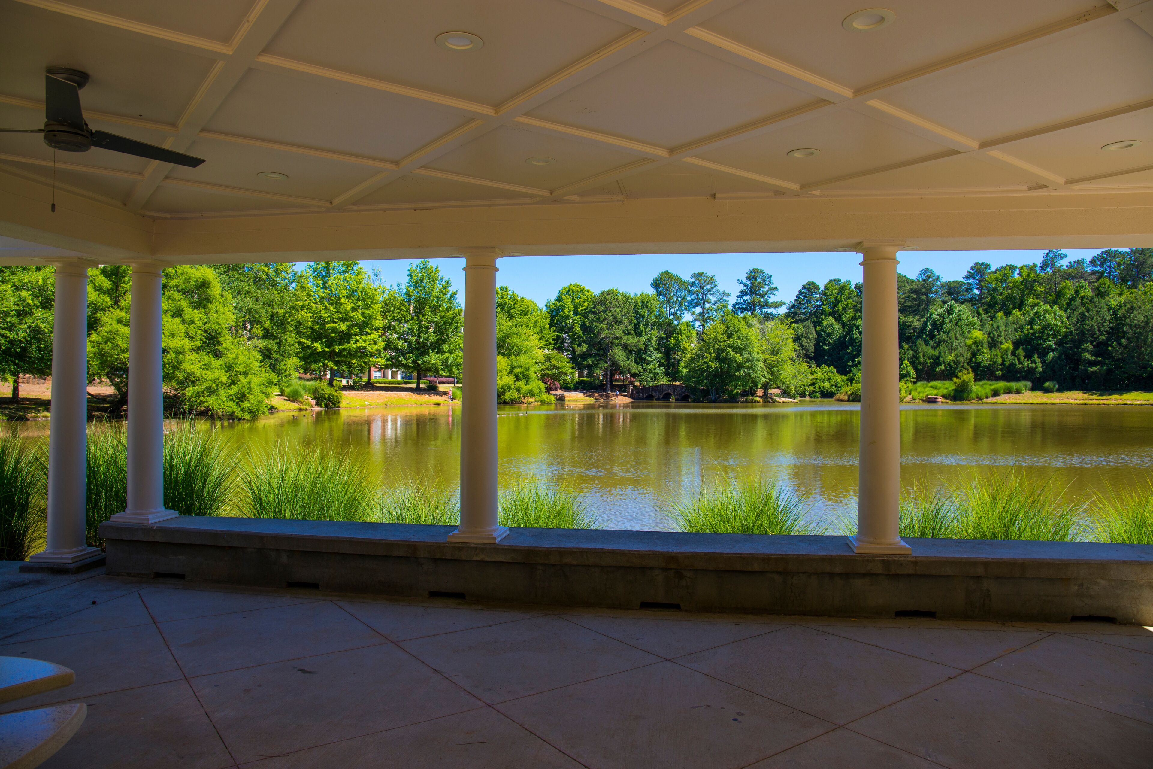 a white pergola with round pillars on the banks of a silky green pond with ceiling fans and round stone park benches surrounded by lush green trees, grass and plants at Mill Creek Pond in Alpharetta