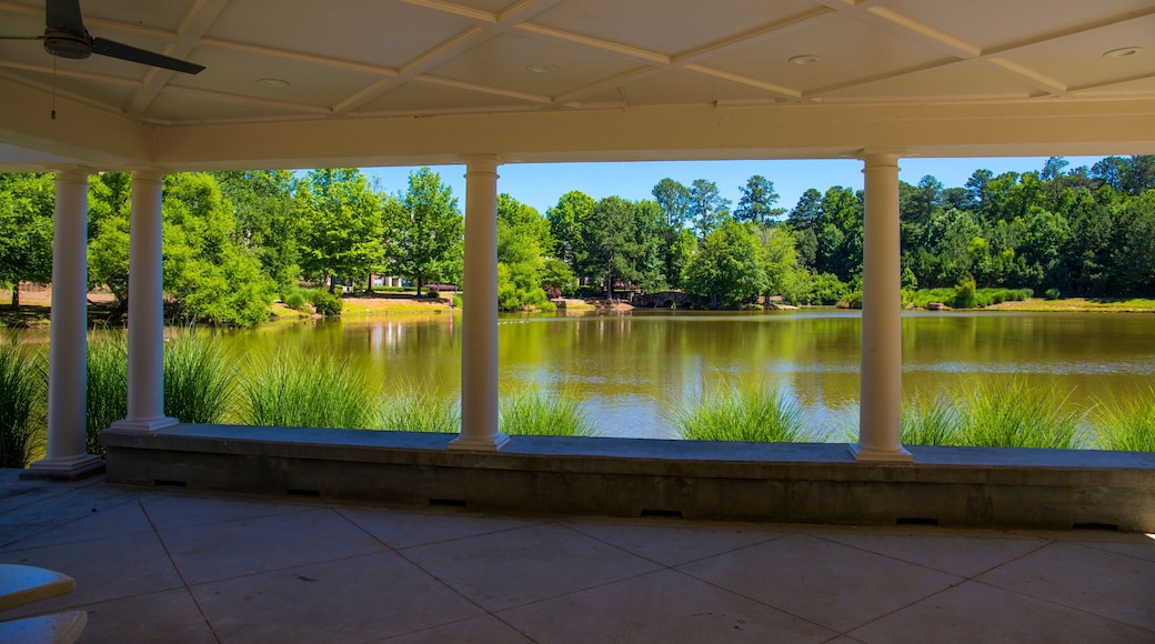 a white pergola with round pillars on the banks of a silky green pond with ceiling fans and round stone park benches surrounded by lush green trees, grass and plants at Mill Creek Pond in Alpharetta