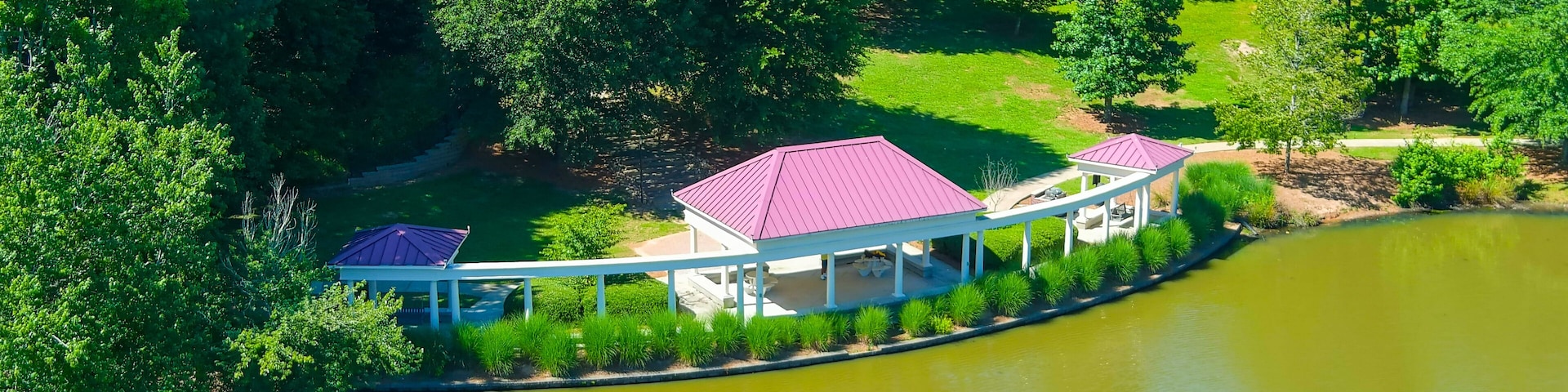 an aerial shot of a gorgeous summer landscape at the pound with silky green water surrounded by lush green trees, grass and plants and white and red pergola on the banks at Mill Creek Pond