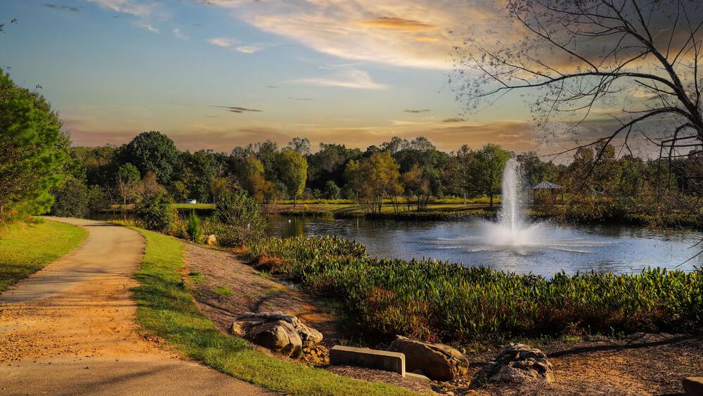 a shot of winding footpath along a lake with a water fountain surrounded by lush green and autumn colored trees and plants with powerful clouds at sunset at Garrard Landing Park in Alpharetta Georgia