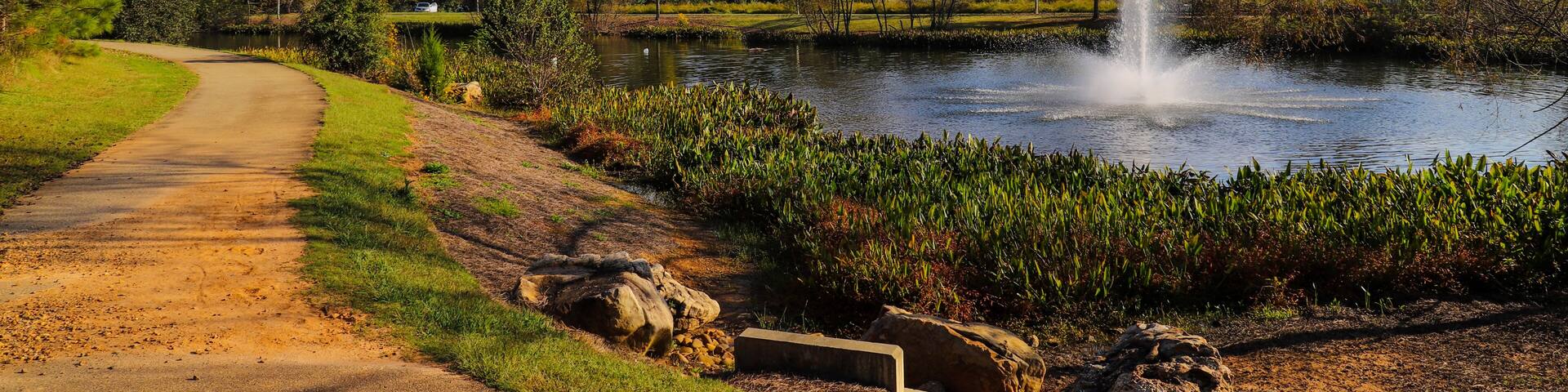 a shot of winding footpath along a lake with a water fountain surrounded by lush green and autumn colored trees and plants with powerful clouds at sunset at Garrard Landing Park in Alpharetta Georgia