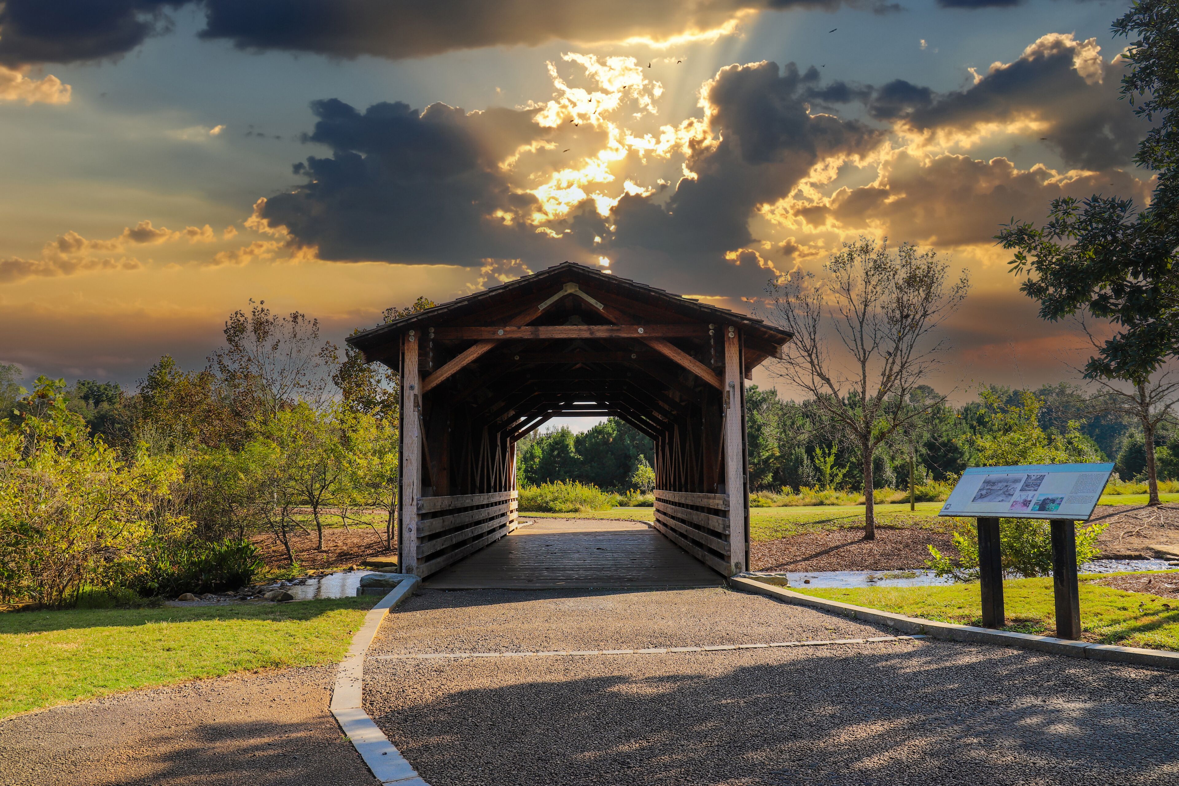 a shot of a wooden covered bridge over a river surrounded by lush green and autumn colored trees with lush green grass and powerful clouds at sunset at Garrard Landing Park in Alpharetta Georgia USA	