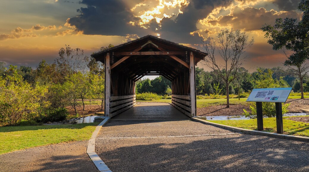 a shot of a wooden covered bridge over a river surrounded by lush green and autumn colored trees with lush green grass and powerful clouds at sunset at Garrard Landing Park in Alpharetta Georgia USA