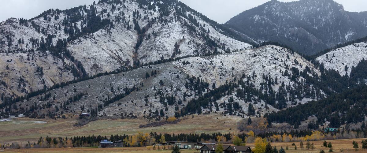 Un paysage montagneux aux sommets enneigés, une vallée parsemée de maisons et des arbres aux couleurs automnales.